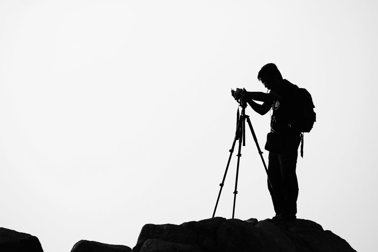 A photographer with a backpack uses a tripod camera on a cliffs edge during a cloudy day.